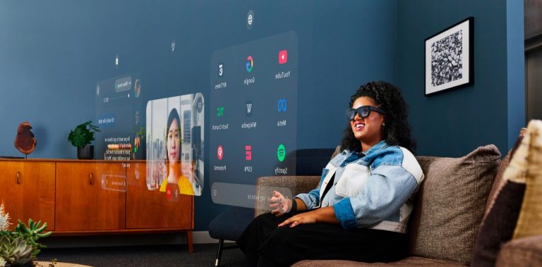 A woman sits on a couch taking a video call and seeing application icons on screens hovering in front of her and Meta's Orion, a product prototype of their yet to be released AR system. Photo courtesy of Meta.